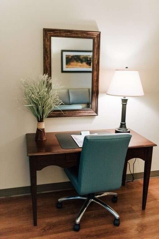 Desk with teal chair, mirror, lamp, and vase of greenery in a room.