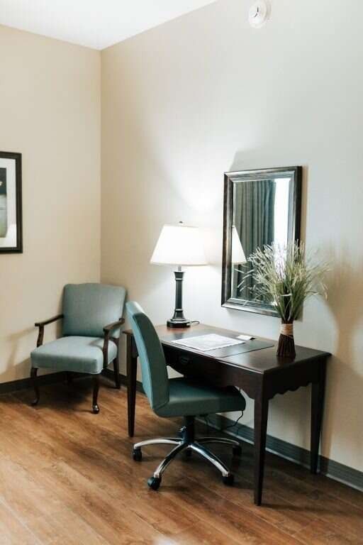 A desk with a chair, lamp, and mirror sits in a corner of a room with a chair nearby.