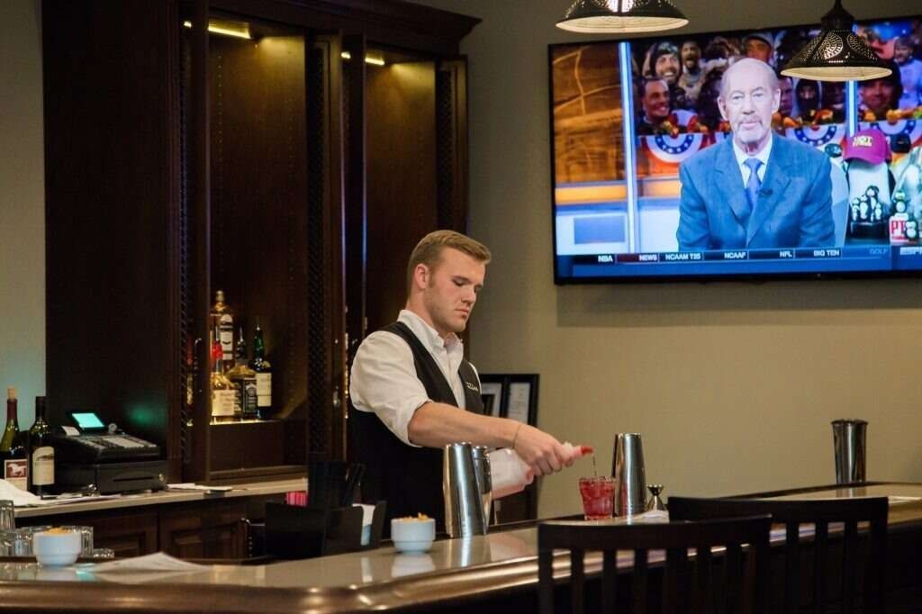 Bartender preparing a cocktail at a bar, TV in the background.