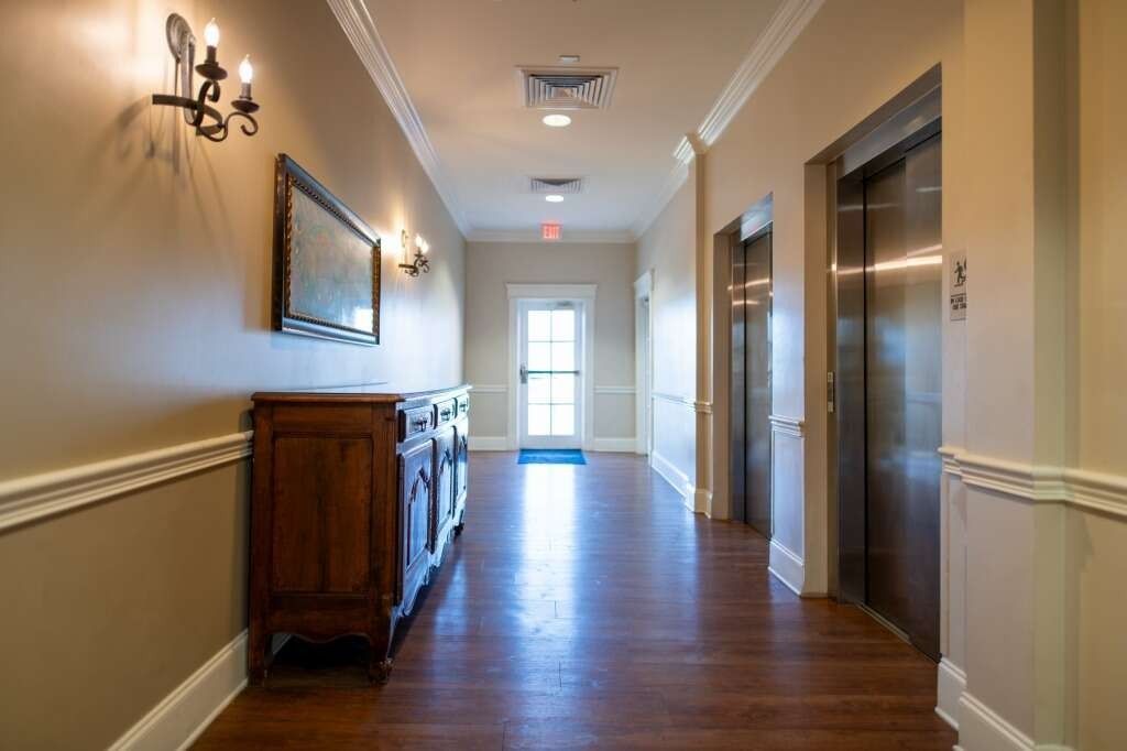 Hallway with elevators, wooden cabinet, sconces, and a door. Beige walls, wood floor.