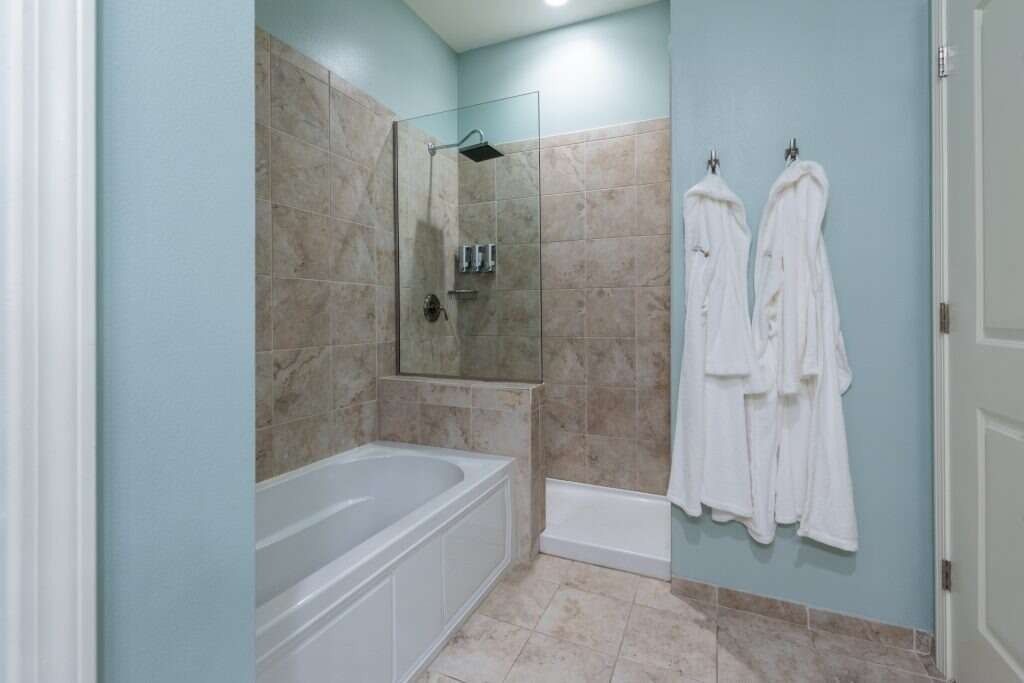 Bathroom with a bathtub, glass shower door, light blue walls, and two white robes hanging on hooks.