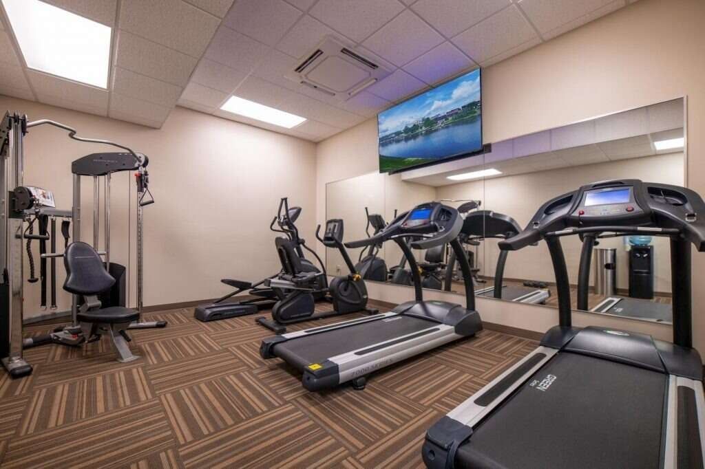 Gym with weight machine, treadmills, elliptical, and a TV on the wall with a mirror, all on a patterned carpet.