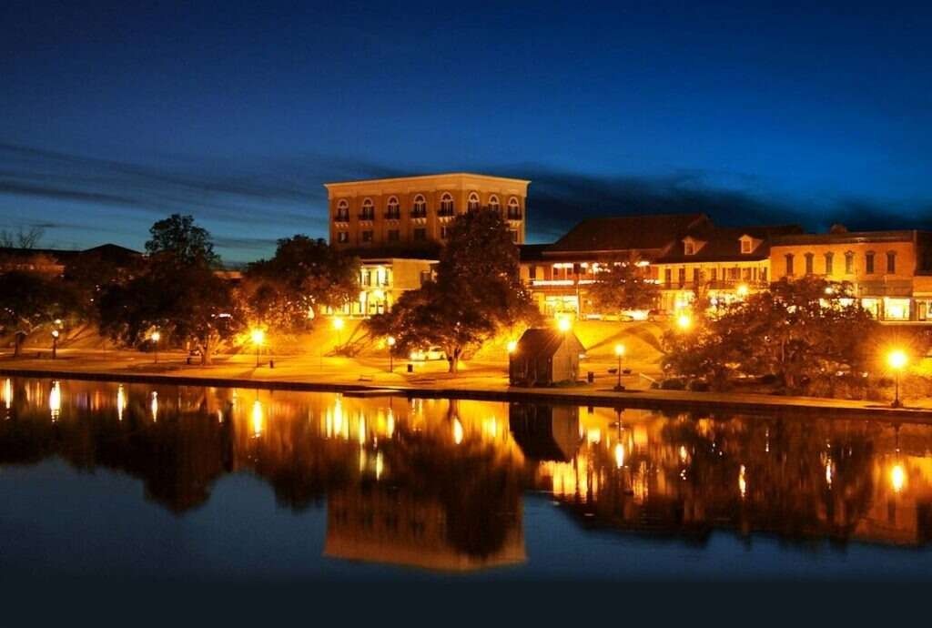Night view of a riverside town with illuminated buildings reflected in calm water under a dark blue sky.