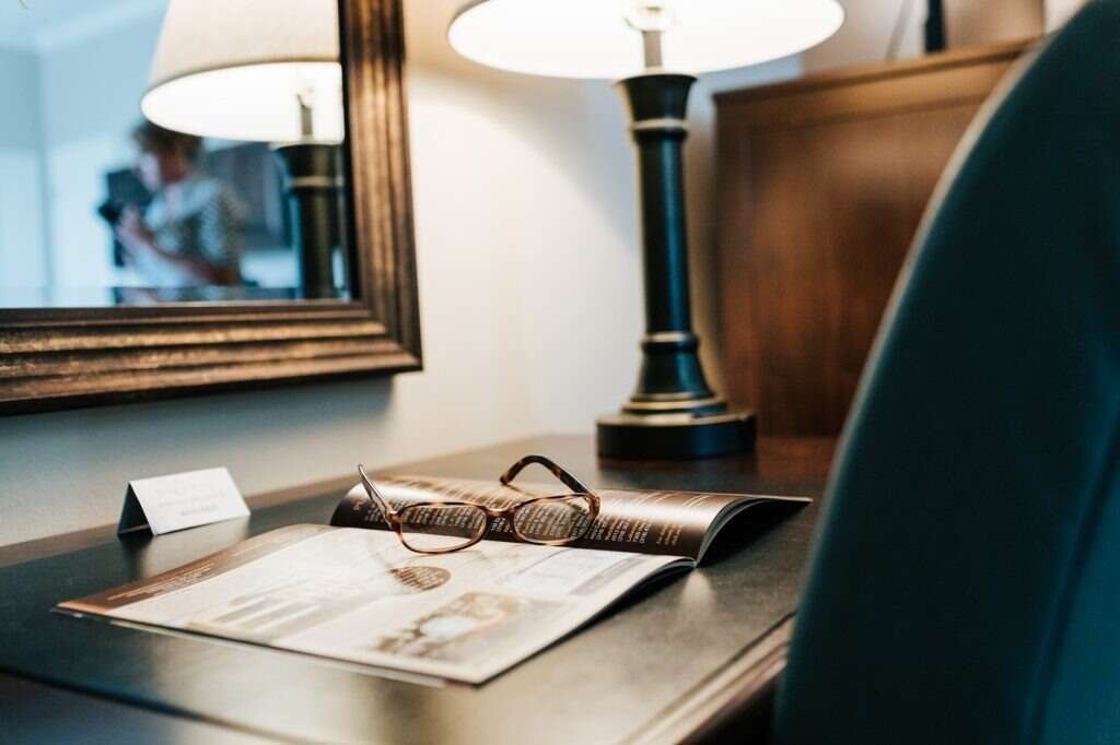 Desk with open magazine, glasses, and lamp. A mirror reflects a person in the background.