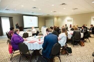 People at tables in a conference room with a presenter, projection screen, and a carpeted floor.